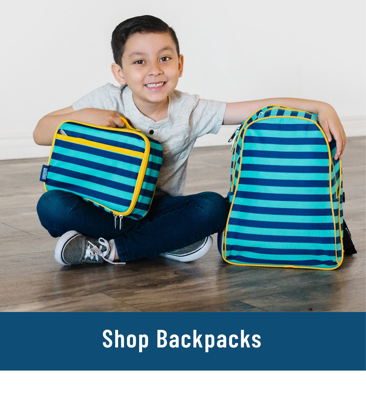Link to shop backpacks. Image of boy sitting on floor with blue stripes backpack and lunch box