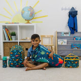 Child sitting indoors with dinosaur-pattern backpack, nap mat, lunch bag, and water bottle arranged around them.