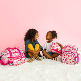 Two girls smiling at each other sitting on fluffy carpet with a backpack, nap mat, duffel bag, and toiletry bag with Strawberry patch pattern.