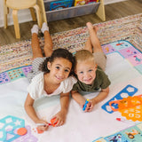 Two young children lying on a colorful doodle mat indoors, drawing and smiling together.